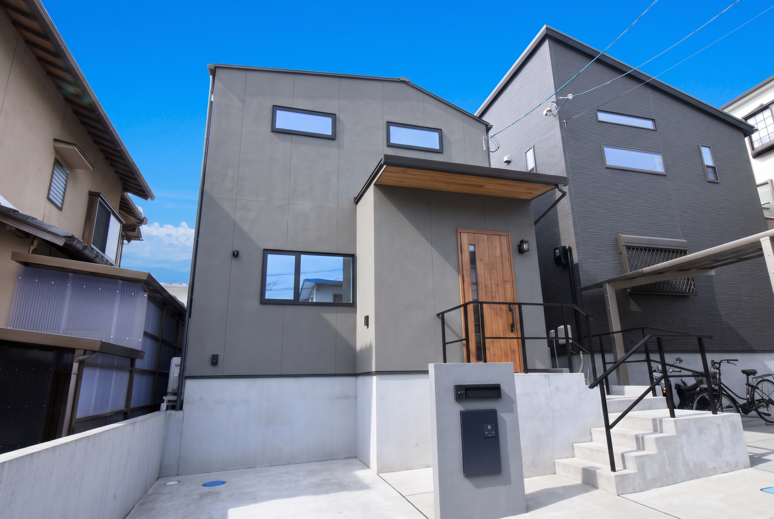 グレーの外壁と片流れ屋根がスタイリッシュなモダンテイストの二階建て住宅の外観。コンクリート打ちっ放しのアプローチ階段に黒いアイアン手すり、木目調の玄関ドアを合わせた洗練されたデザインです
