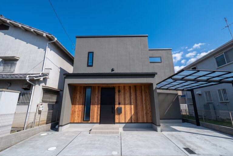 グレーの外壁とスクエアなフォルムが特徴的なモダンスタイルの住宅外観。玄関周りに施された木目調のアクセントと黒いドアが調和し、右側にはカーポートを備えています。