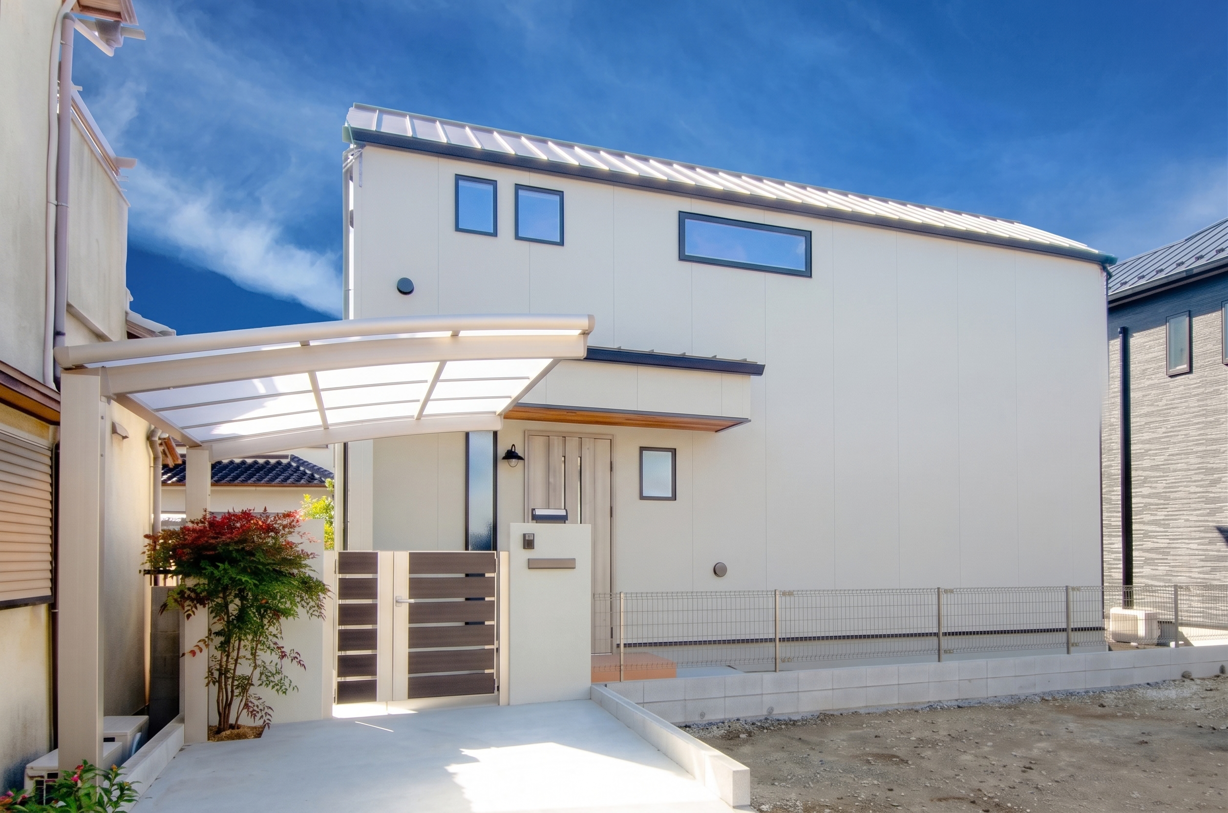 青空に映える白い外壁とシャープな片流れ屋根が特徴的な、シンプルモダンな住宅の外観。手前にはカーポートと木目調の門扉を備えた駐車スペースがあります。