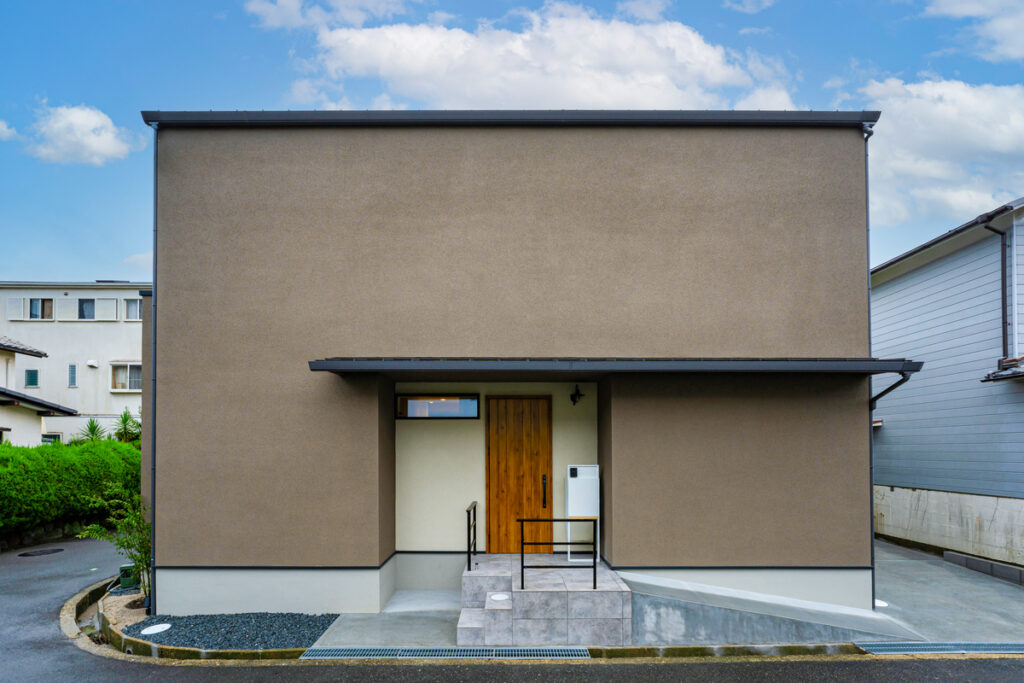 塗り壁の質感と木目調のドアが調和した、窓を最小限に抑えたモダンでシンプルなボックス型の外観。