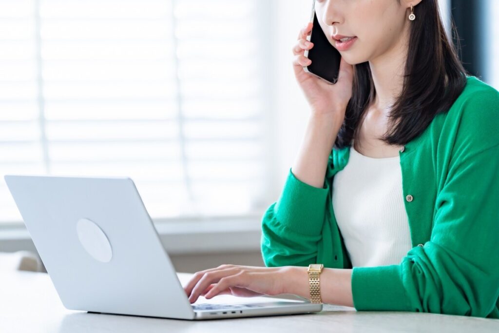 パソコンを操作しながらスマートフォンで通話をする、緑色のカーディガンを着た女性