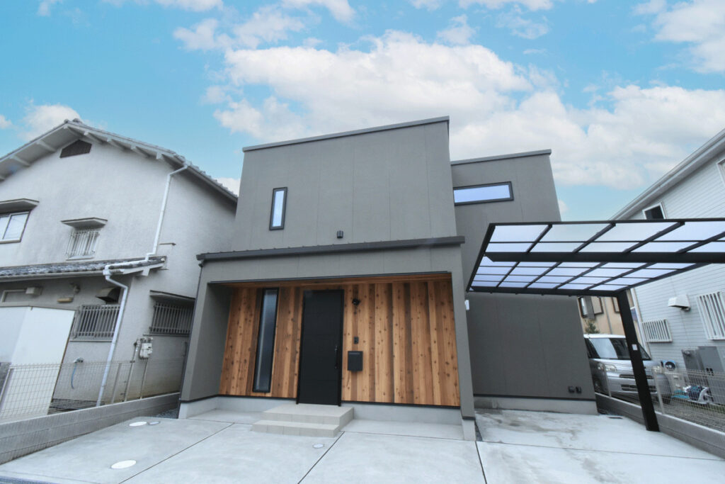 グレーの外壁と玄関周りの温かみのある板張り、黒のサッシが調和したスタイリッシュなボックス型のモダン住宅。