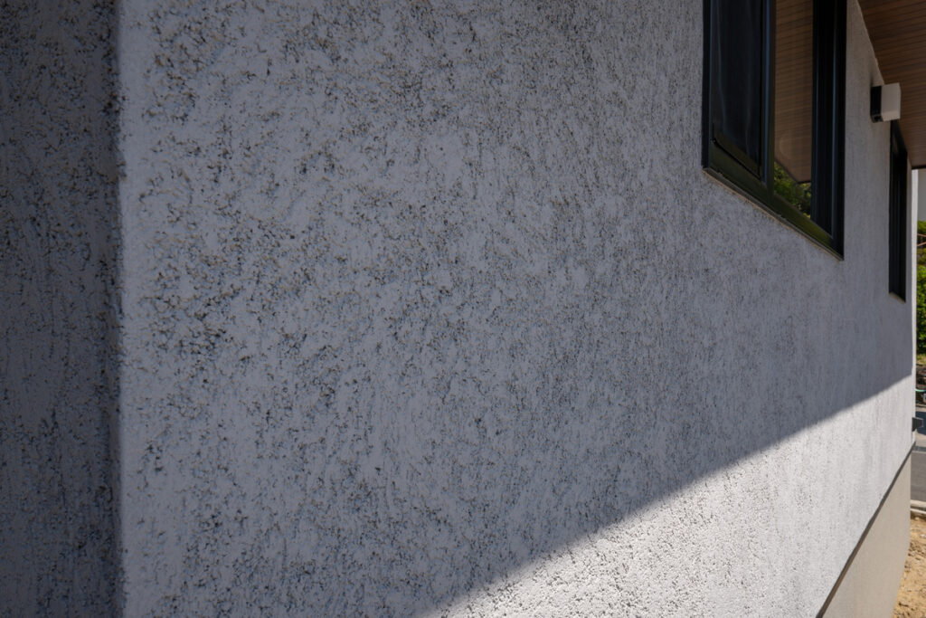 職人の手仕事を感じさせる、豊かな風合いの塗り壁外壁のディテール。太陽の光によって浮かび上がる細かな陰影と窓枠のコントラスト