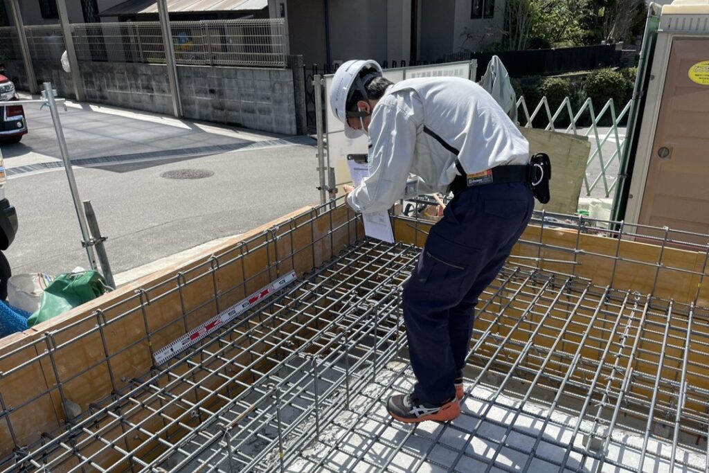 建物の強度を左右する基礎工事現場で、現場監督がスケールを用いて鉄筋の間隔や配置を厳格に確認する配筋検査の様子。