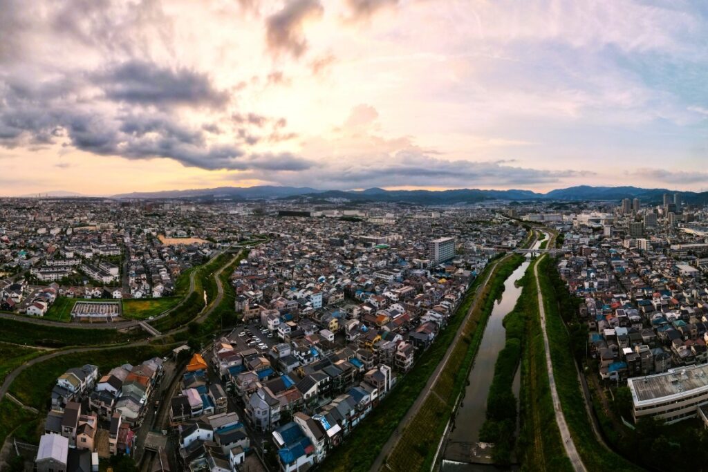 川の流れに沿って住宅街が広がる高槻市周辺の街並みと、遠くに北摂の山々を望む夕暮れ時のパノラマ風景。
