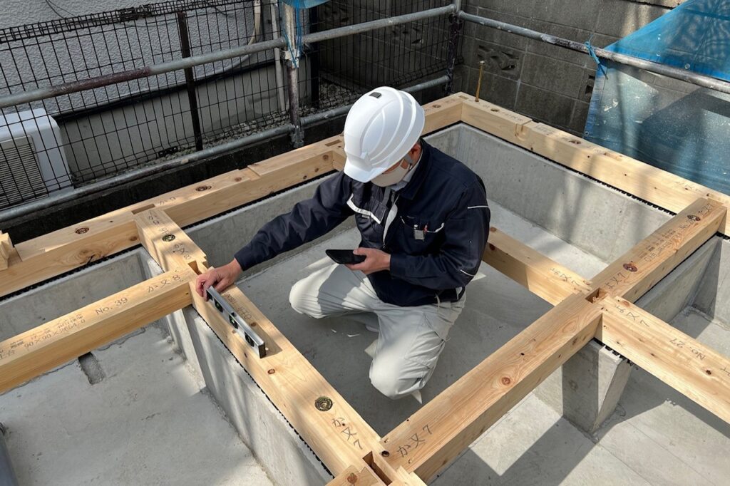 コンクリート基礎の上に組まれた木造住宅の土台で、現場監督が水平器を用いて施工精度を検査する様子。