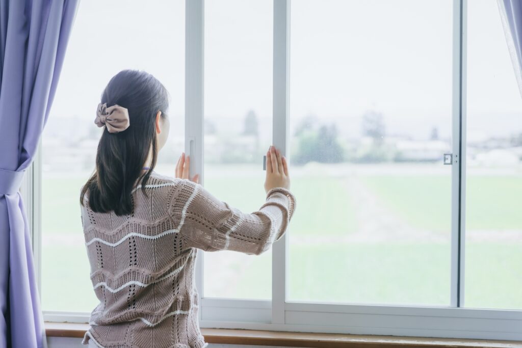 白いサッシの窓を開けて外の景色を眺める女性の背中。自然光が差し込む明るい室内と、窓辺を彩る落ち着いた紫色のカーテン