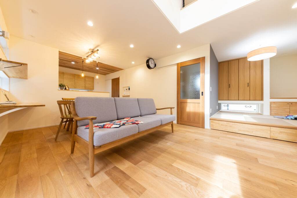 吹き抜けから光が降り注ぐ、無垢の床が心地よいリビング。北欧風のソファを主役に、奥にはキッチン、右側には小上がりの畳コーナーが広がる。