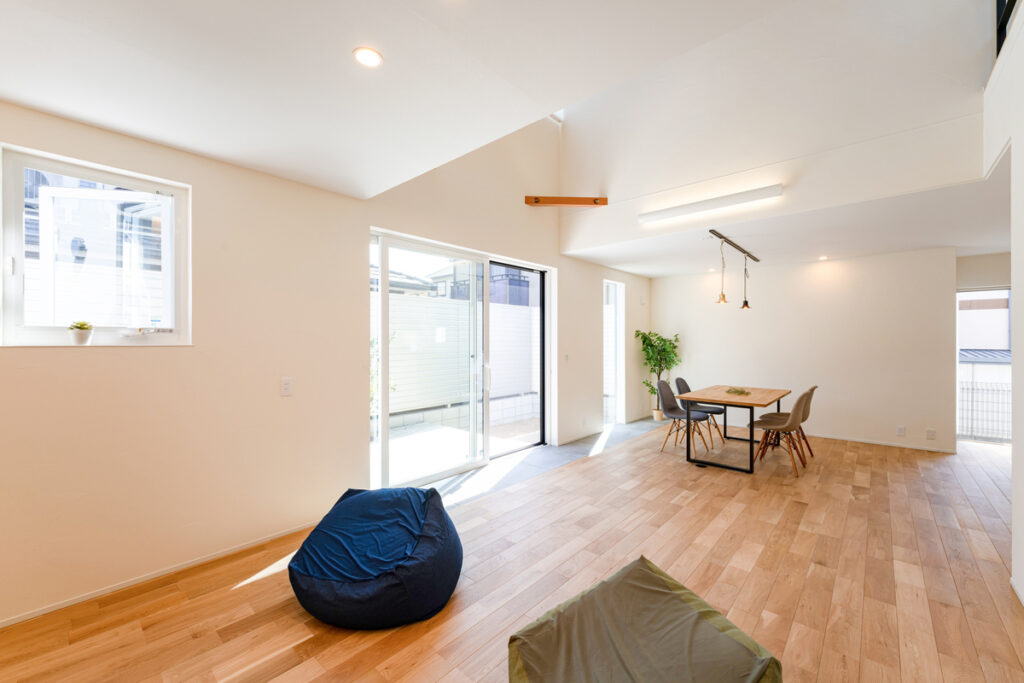 吹き抜けが開放的な、白い壁と無垢床のナチュラルなリビング。大きな掃き出し窓から明るい光が差し込み、テラスへと視線が抜ける明るい住空間。