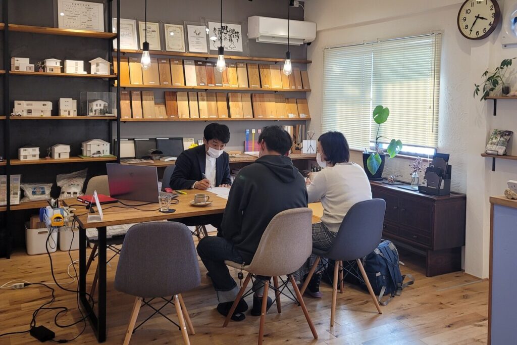 木材サンプルや住宅模型が並ぶショールームで、スタッフと施主が家づくりの打ち合わせを行っている風景。