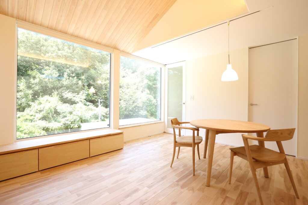 勾配天井の板張りと大きな窓が開放的なダイニング。木の質感を活かした円形テーブルと椅子が並び、外の緑と光を取り込む温かみのある住空間。