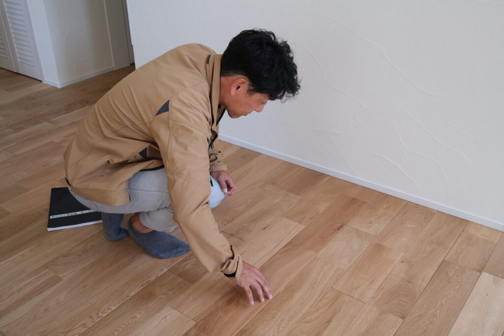 施工されたばかりの新しい無垢床に触れ、その質感や仕上がりを確認する男性。木の温もりや肌触りの良さを象徴するシーン。