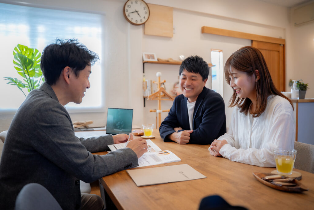 明るい商談スペースにて、カタログを広げながら笑顔で家づくりの相談を進めるご夫婦と、それに応える工務店スタッフの対面打ち合わせ。