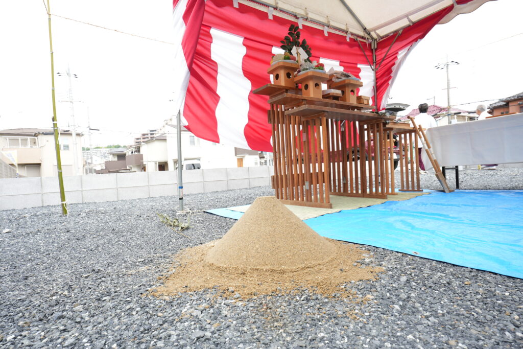 住宅建築の安全を祈願する地鎮祭の様子。手前に円錐形に整えられた盛り砂があり、奥の紅白幕を張ったテント内には、お供え物が並ぶ伝統的な祭壇が設営されています。