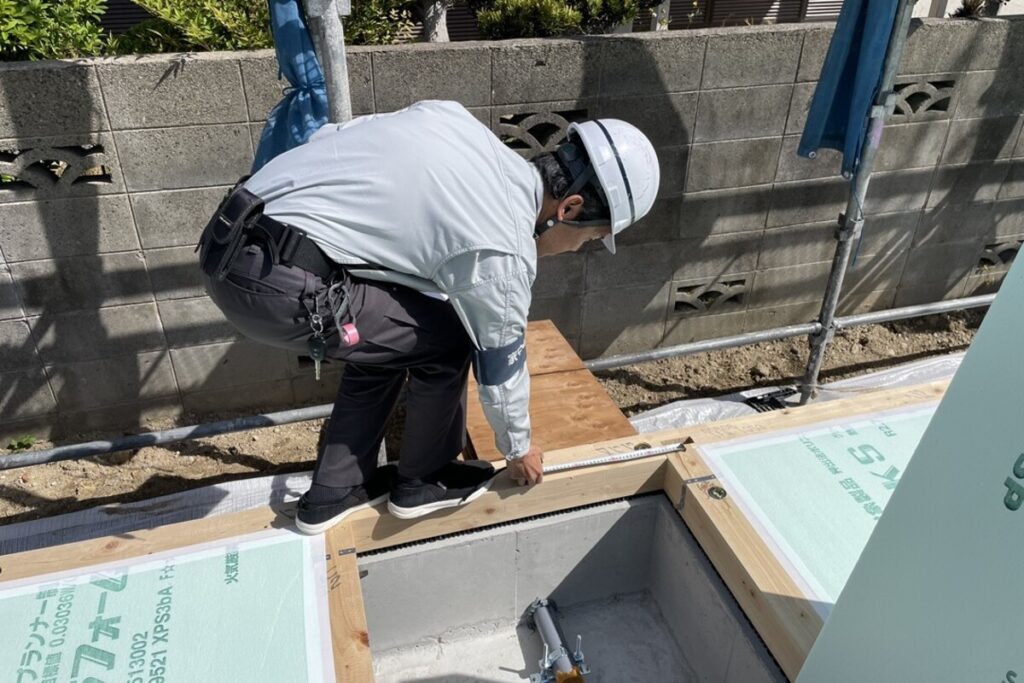 床下断熱の施工において、断熱材の設置状況や土台との接合部をメジャーで厳密にチェックし、住宅の基本性能を保証する現場管理の様子。