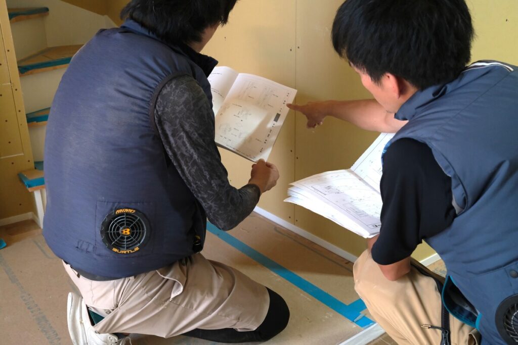 施工現場にて、空調服を着用した職人が図面と照らし合わせながら細部の収まりを確認する、高品質な施工を実現するための入念な打ち合わせ。