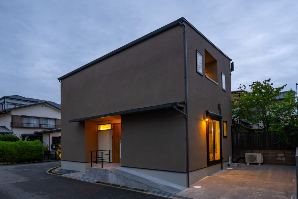 夕暮れ時に室内からの温かい光が窓から漏れる、落ち着いたブラウンの塗り壁が美しいモダンな外観。玄関へは緩やかなスロープが続くバリアフリー設計。