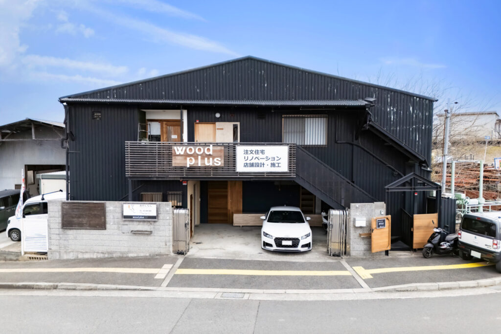 黒いガルバリウムの外壁に「woodplus 注文住宅 リノベーション」の看板が掲げられたオフィス・店舗の外観