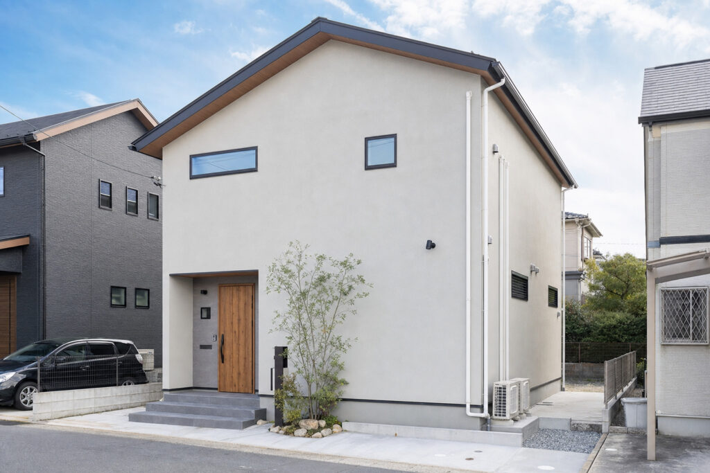 白い外壁と木目調の玄関ドアが特徴的なモダンな二階建て住宅の外観。側面にはエアコンの室外機が2台並んで設置されています。
