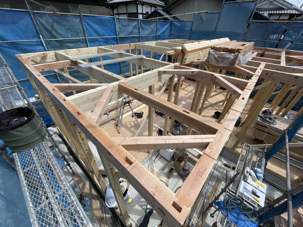 足場の上部から見下ろした、建築中の木造住宅の骨組み。柱や梁、屋根を支えるための小屋組みが組まれており、下方の床面では複数の作業員が作業をしています。周囲は青い飛散防止ネットを張った足場で囲まれています。