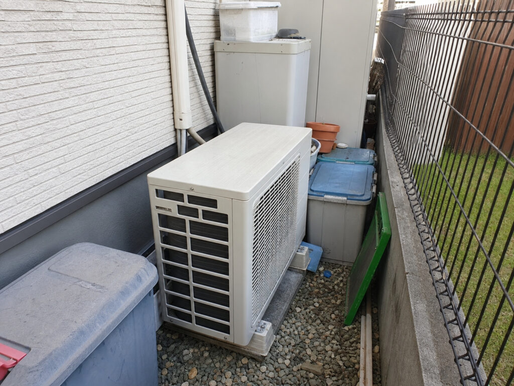 住宅の外壁と境界フェンスの間の、砂利が敷かれた狭い犬走りスペースに設置されたエアコン室外機。
