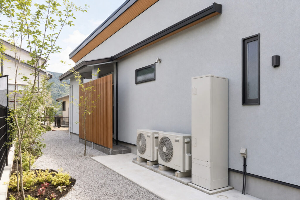 グレーの外壁の住宅側面に並んで設置されたエコキュートの貯湯タンクと2台のエアコン室外機。玄関ポーチの木製目隠しルーバーも見えます。