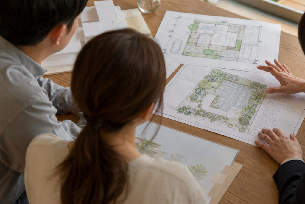 木目のテーブルに住宅の配置図や平面図を広げ、建築士がご夫婦に向けてプランの説明をしている打ち合わせの様子。