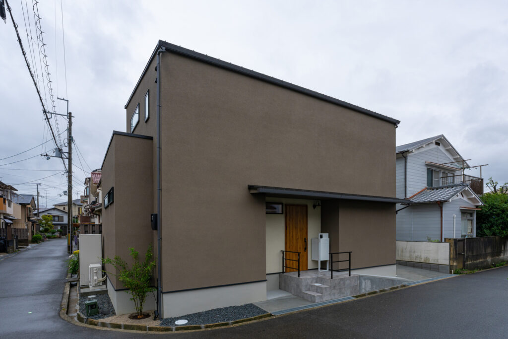 ダークブラウンの落ち着いた外壁が特徴的な、キューブ型のモダンな二階建て住宅の外観。木製玄関ドアや黒いアイアン手すりがアクセントになっており、側面には室外機とエコキュートが配置されています。