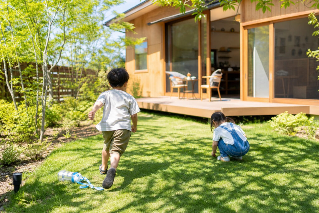 木の温もりを感じる板張りの外壁と広いウッドデッキがある住宅。緑豊かな芝生の庭で遊ぶ子どもたち。