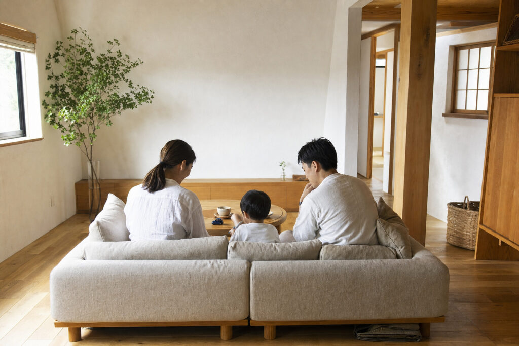 無垢材の床と白い塗り壁に囲まれたナチュラルなリビング。ローソファに座り、家族3人でくつろいでいる様子です。