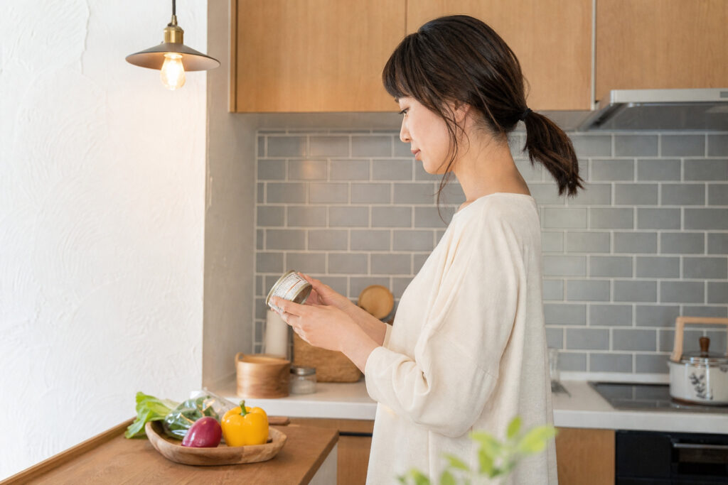 木目調のキャビネットとグレーのタイル張りの壁が特徴的なキッチンで、備蓄用缶詰の表示を確認する女性。