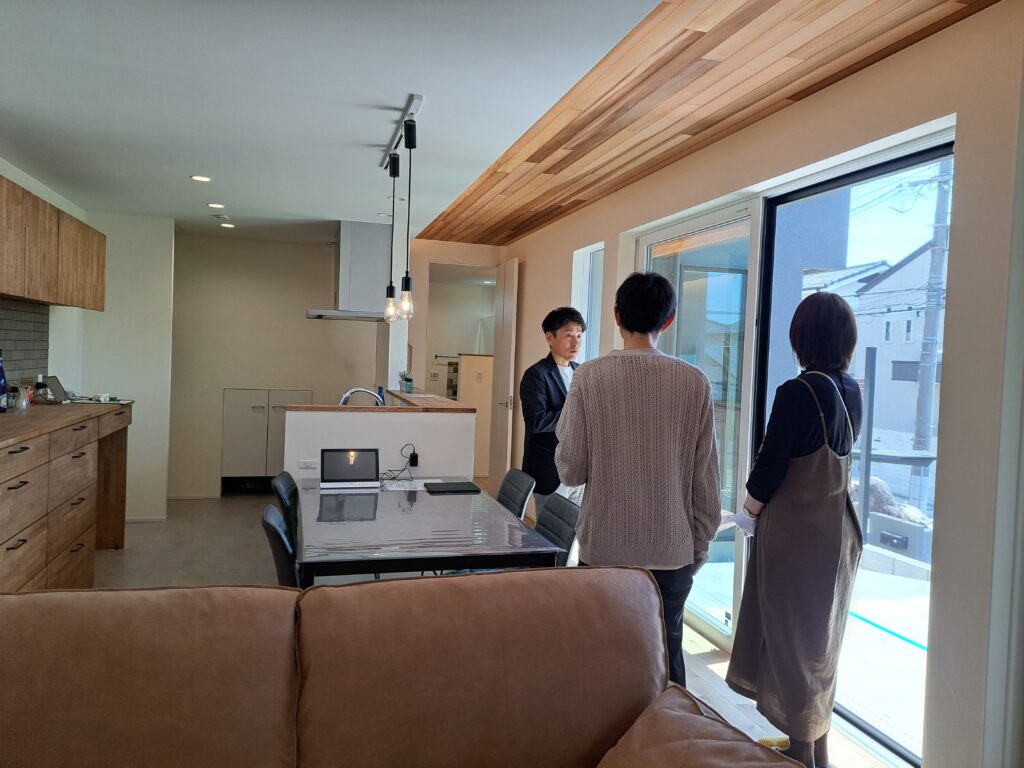 板張りの天井や木目の背面収納が温もりを演出するLDK。大きな掃き出し窓のそばで、担当者の説明を聞きながら見学する夫婦。