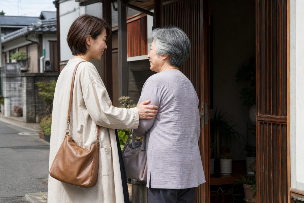 趣のある木製の引き戸の玄関先で、訪問してきた若い女性を笑顔で出迎えるシニア女性の様子。