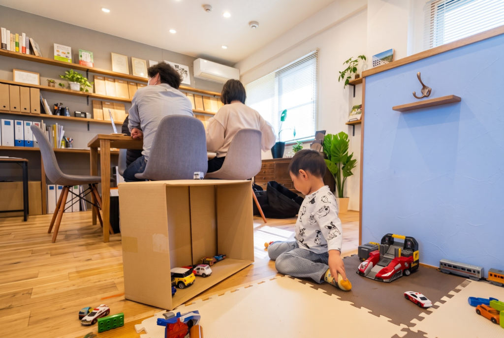 木目の床と壁面のオープン棚、水色のパーティションがある打ち合わせスペース。奥のテーブルでご夫婦が相談する間、手前のマット上で子どもがおもちゃで遊んでいます。