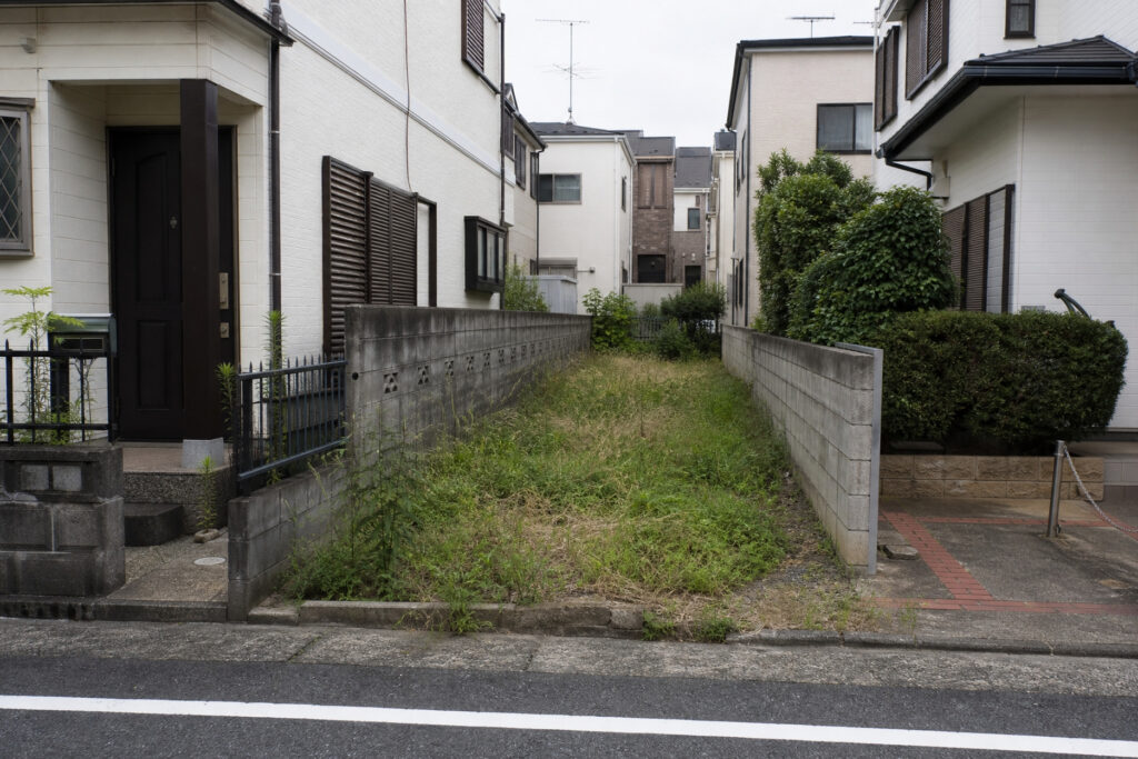 住宅街に位置する、ブロック塀で区切られた細長い空き地。両隣に家が密集する狭小地での土地探しの状況を示しています。