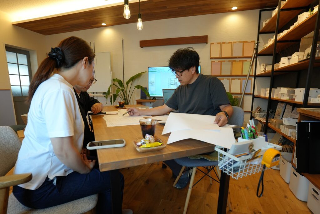 建築模型や素材サンプルが並ぶ打ち合わせスペースで、設計士から図面の詳細な説明を受ける夫婦。