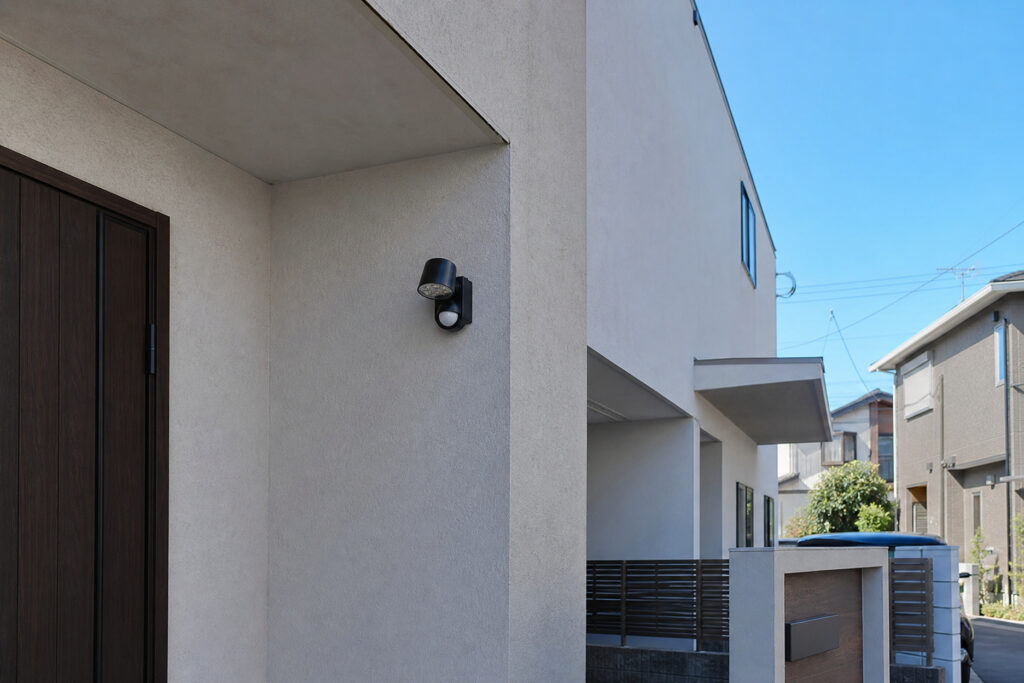 白い外壁に設置されたセンサー付き屋外照明と、木目調の玄関ドアが見える住宅の外観。