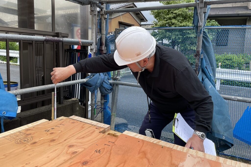 足場が組まれた建築現場で、木質パネルの施工箇所にメジャーを当てて正確に寸法を確認している現場担当者。