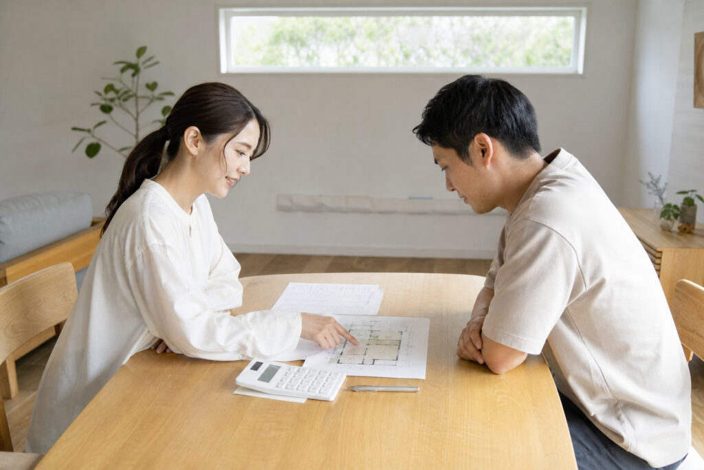 明るい室内で無垢材のテーブルを囲み、図面を指し示しながら間取りや資金計画の相談をしている夫婦の風景。