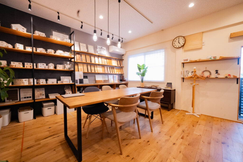 工務店の打ち合わせスペース。無垢材の床やテーブルが配され、壁面の棚には多数の住宅模型や建材サンプルが展示されています。