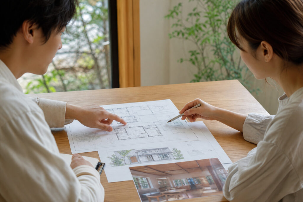 木のテーブルに広げた間取り図や外観パースを見ながら、家づくりの打ち合わせをしている様子。