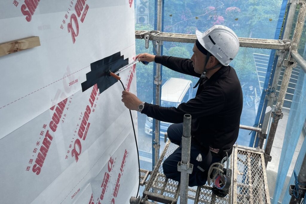 建築現場の足場上で外壁の透湿防水シートを施工する作業員。配管の貫通部周りに防水テープを貼り、雨水の侵入を防ぐ処理を行っています。