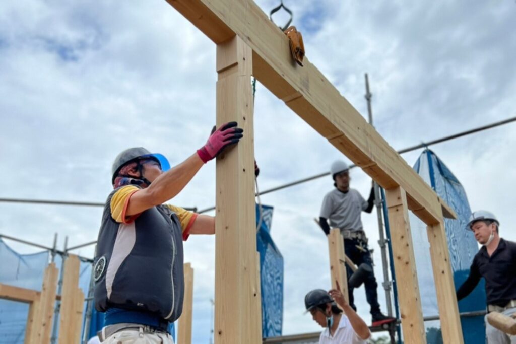 青空の下、木造住宅の建築現場で大工が太い無垢材の柱と梁を丁寧に組み上げている上棟作業の様子。