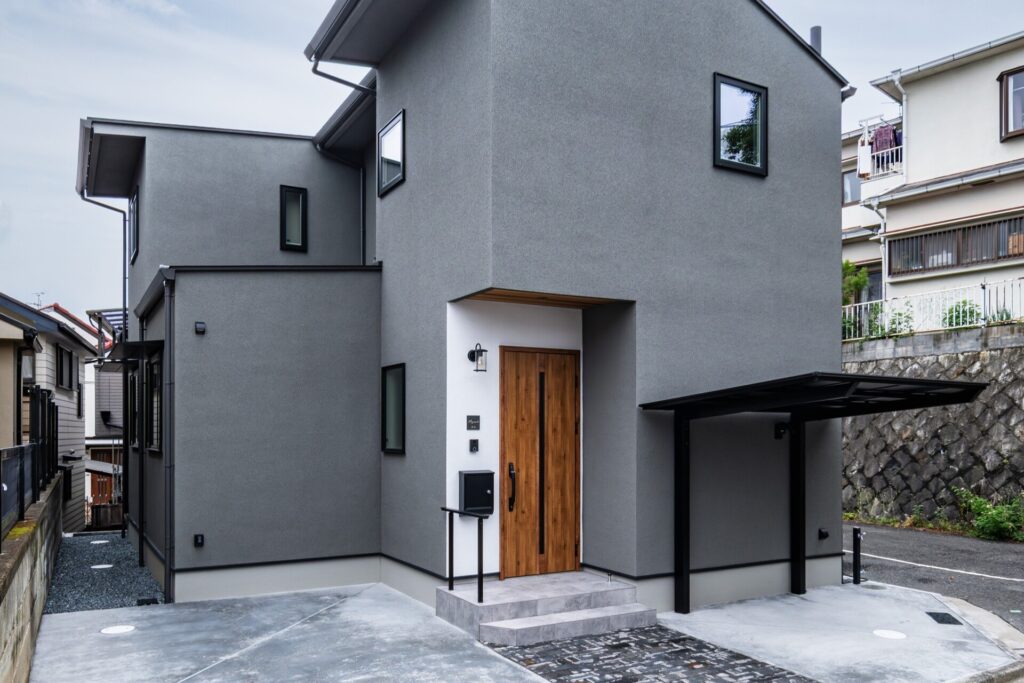グレーの外壁と片流れ屋根が特徴的なモダンスタイルの住宅外観。木目調の玄関ドアと黒いカーポートがアクセントになっています。