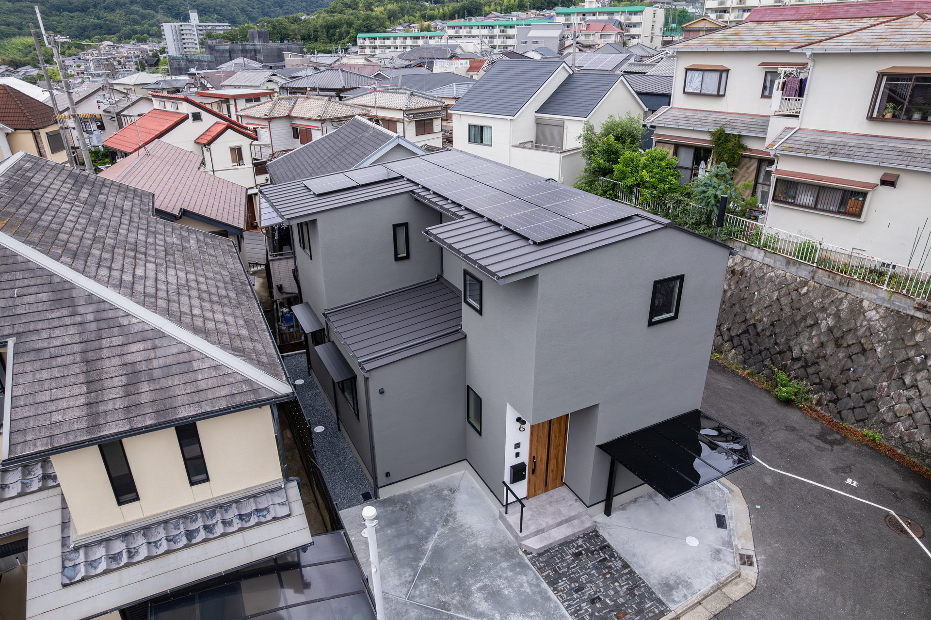 住宅密集地に建つ、グレーの外壁と太陽光パネルを搭載した金属屋根が特徴的なモダンテイストの住宅外観。