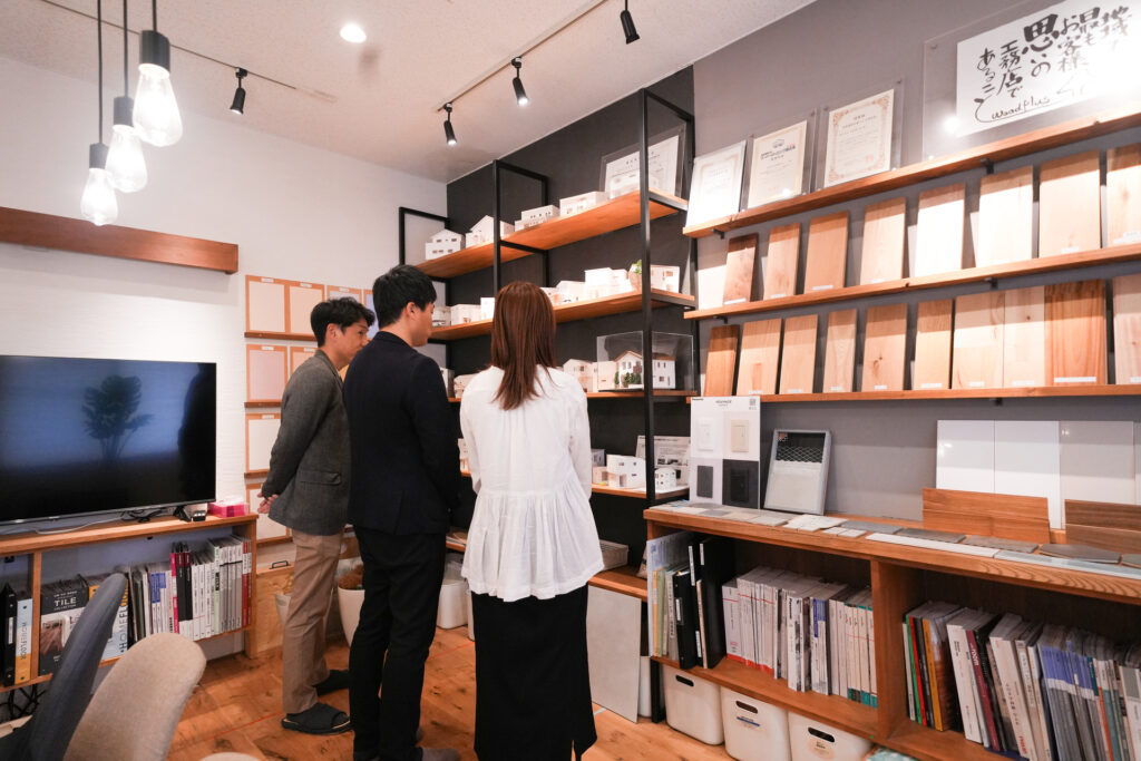 工務店のショールームで、壁面に並んだ無垢材の床材サンプルや住宅模型を見比べながら、素材選びの打ち合わせをしているお客様と担当者。