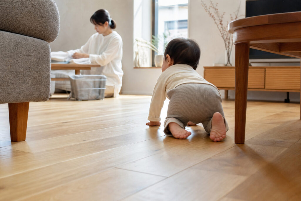 木目が美しい無垢材風フローリングのリビングをハイハイする赤ちゃん。奥には自然光を取り込む窓辺で家事をする母親の姿が見える、暮らしの風景。