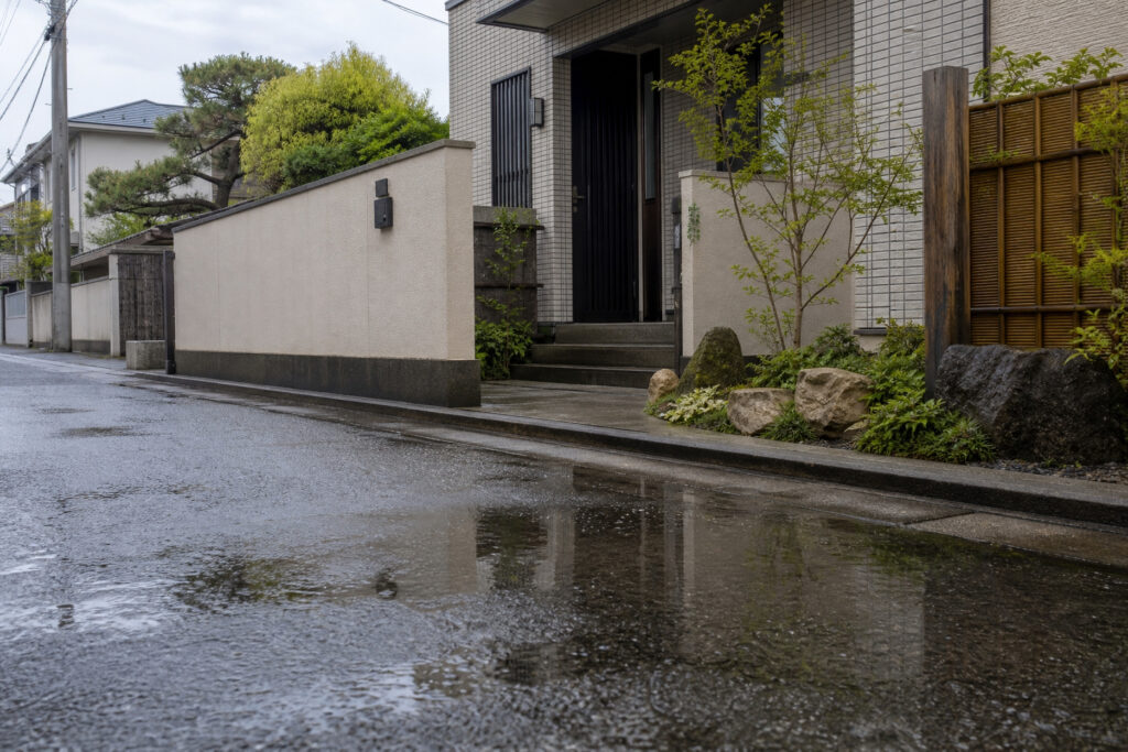 雨上がりの水たまりに反射する和モダンな住宅の外観。白い壁と木の格子、石組みと植栽が特徴的なアプローチ。