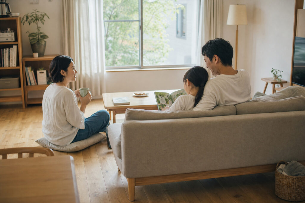 無垢材の床と木の温もりを感じる家具が配置された明るいリビングで、ソファに座り家族でくつろぐ様子。