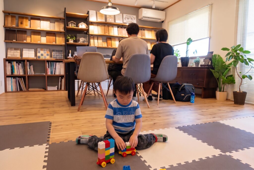 無垢材の床が広がる打ち合わせスペース。手前のキッズスペースで子供がおもちゃで遊び、奥では大人たちが木材のサンプルを前に家づくりの相談をしています。